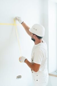 Construction worker using tape to prepare wall surface, wearing safety helmet and gloves.